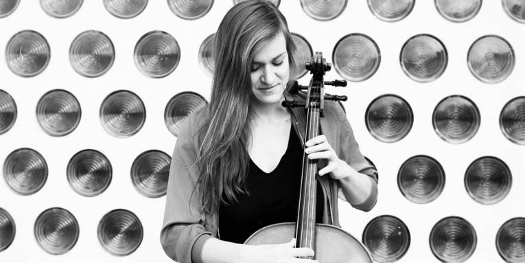 eine junge Frau steht mit ihrem Cello vor einer Wand mit runden Glasbausteinen.